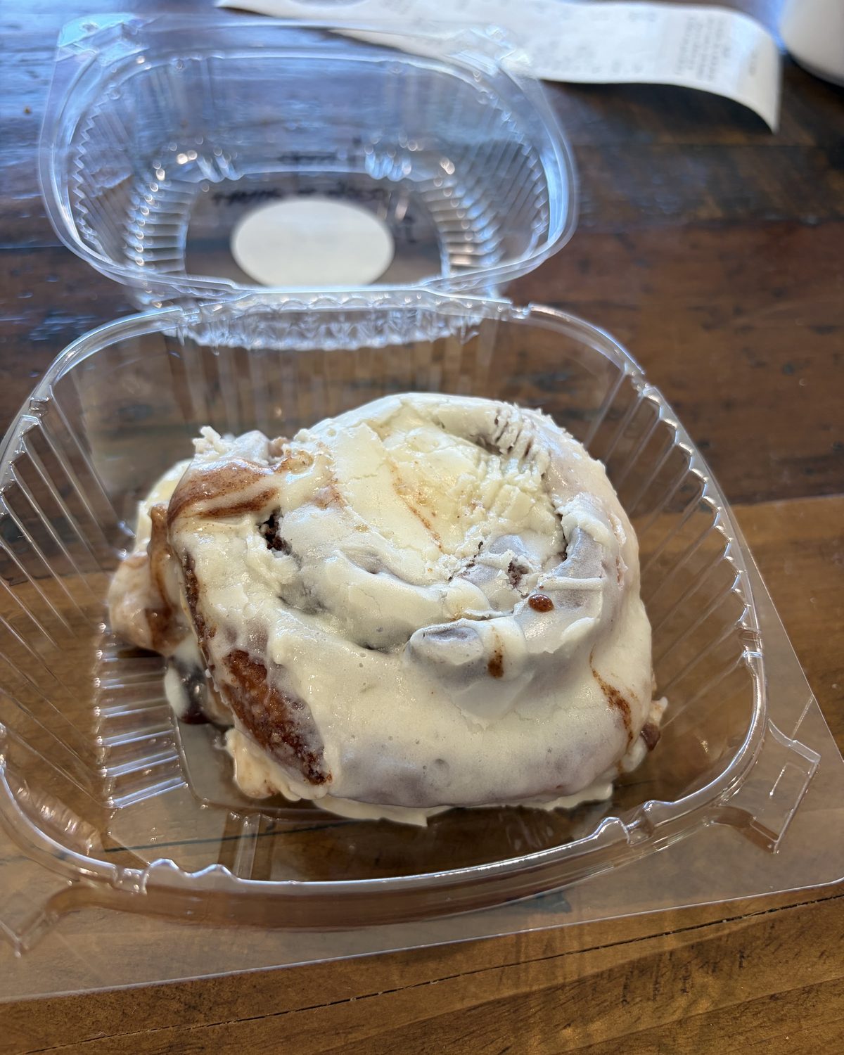 A large iced cinnamon roll in a to-go container on a wooden table
